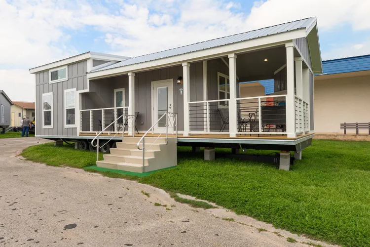 A tiny home sits on blocks on a patch of grass. Stairs lead up to the wrap around porch with seating area and fireplace.