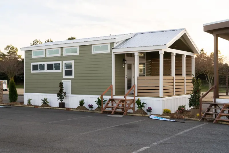 A tiny home sits on a parking lot with plants surrounding it. There is a stairway leading to the front door on the small front porch.